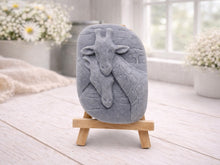 Load image into Gallery viewer, A rectangular handmade vegan soap in a natural tone, featuring a detailed relief of a mother and baby giraffe with their necks intertwined, displayed on a miniature wooden easel against a white wood-panelled background.
