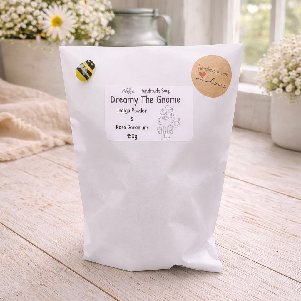 White bag of handmade soap labeled 'Dreamy The Gnome' on a wooden surface with flowers in the background.