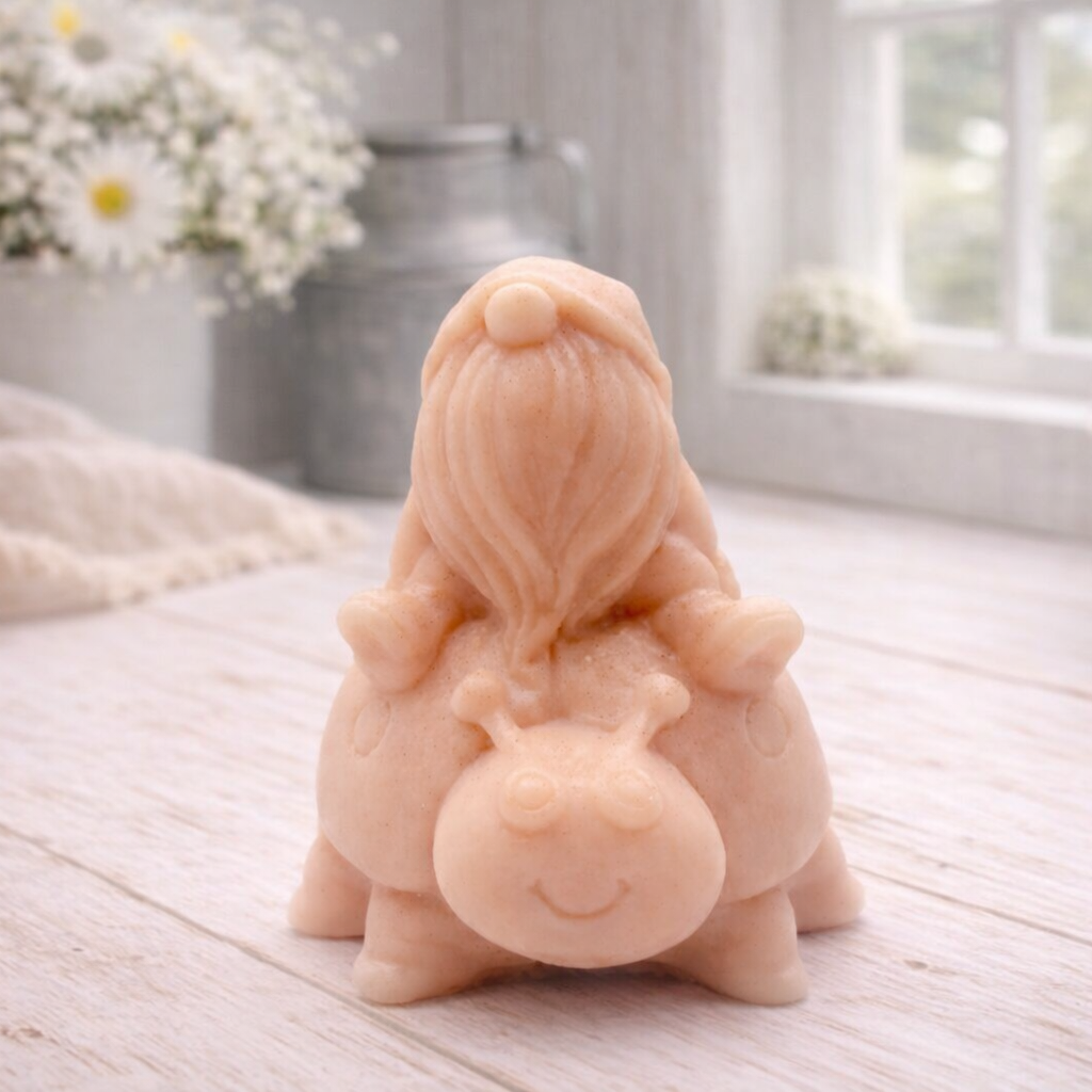 A handmade vegan soap in a warm terracotta-peach tone, sculpted as a whimsical gnome (gonk) with a long, flowing beard sitting on the back of a large, smiling ladybird with visible spots, displayed on a light wooden surface.