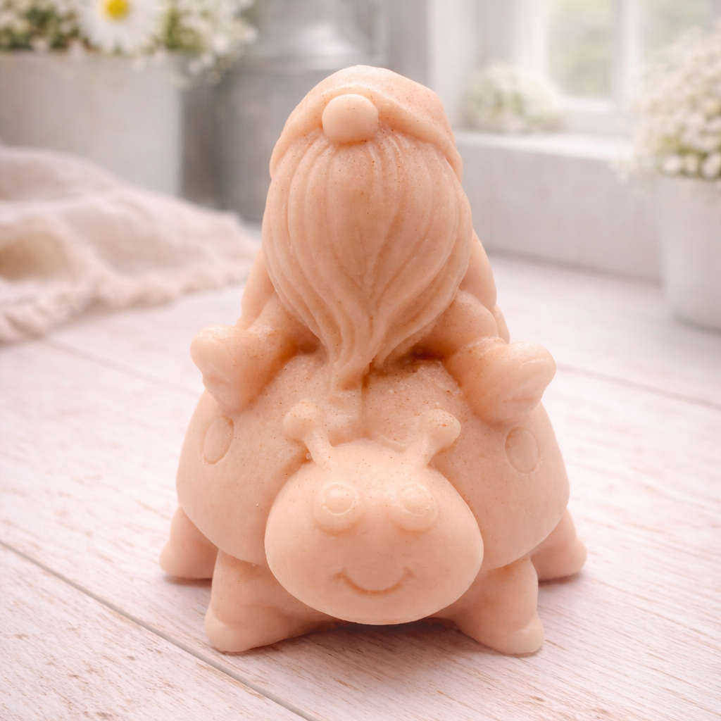 A handmade vegan soap in a warm terracotta-peach tone, sculpted as a whimsical gnome (gonk) with a long, flowing beard sitting on the back of a large, smiling ladybird with visible spots, displayed on a light wooden surface.