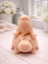 Load image into Gallery viewer, A handmade vegan soap in a warm terracotta-peach tone, sculpted as a whimsical gnome (gonk) with a long, flowing beard sitting on the back of a large, smiling ladybird with visible spots, displayed on a light wooden surface.
