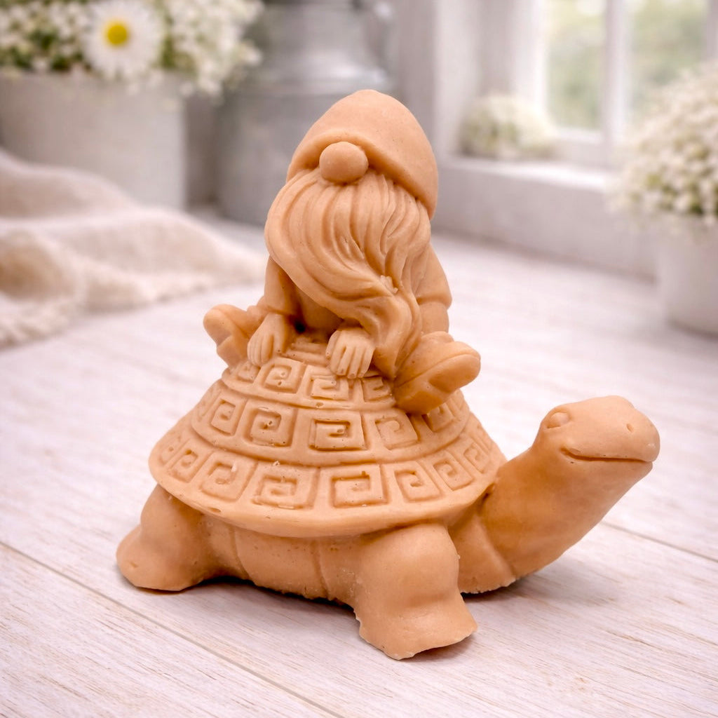 A handmade vegan soap in a natural tone, sculpted as a whimsical gnome (gonk) with a long beard sitting on the back of a large, smiling turtle with a patterned geometric shell.