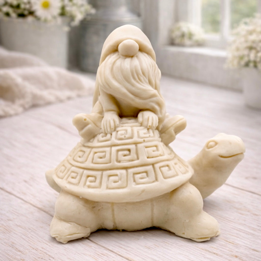 A handmade vegan soap in a natural tone, sculpted as a whimsical gnome (gonk) with a long beard sitting on the back of a large, smiling turtle with a patterned geometric shell.