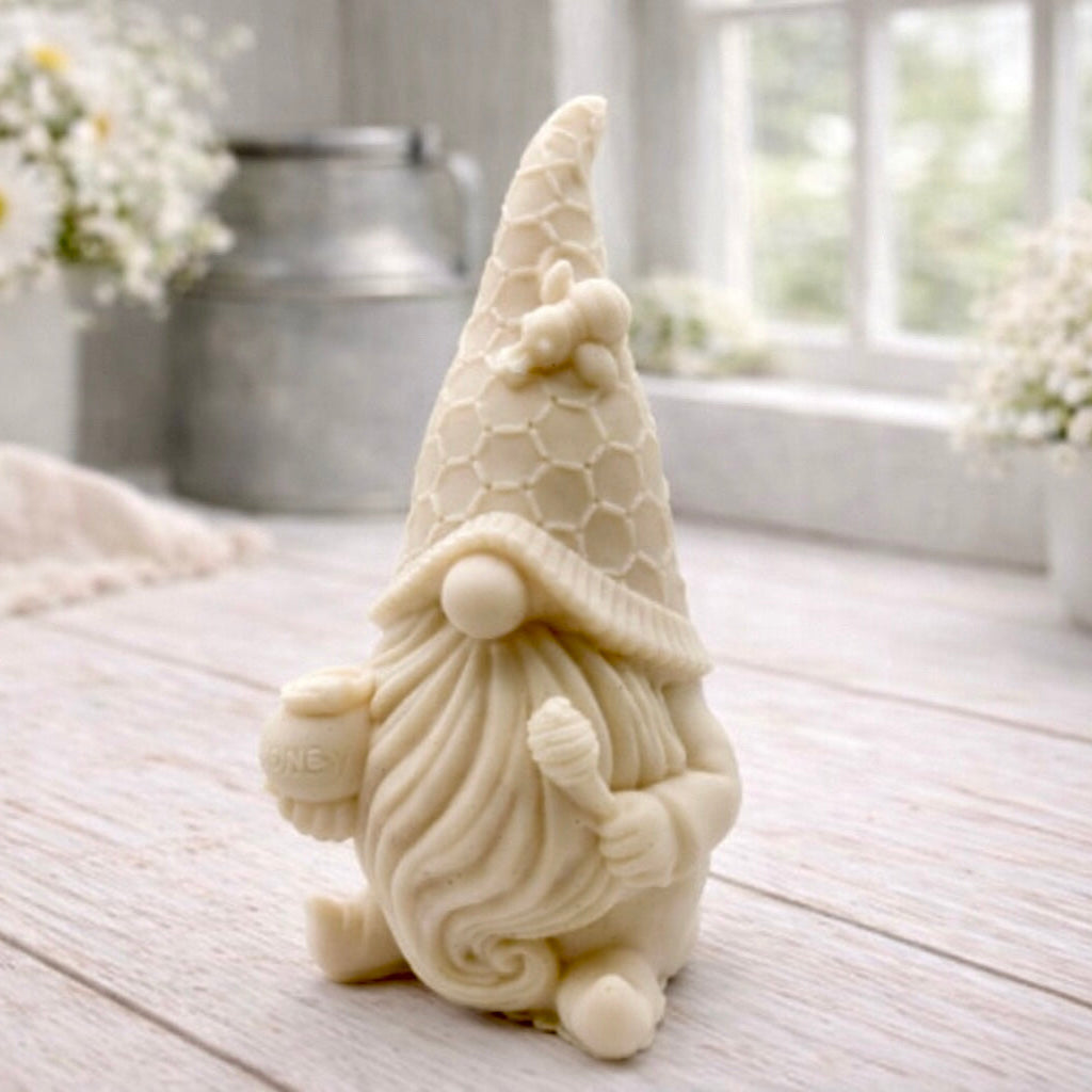 A handmade vegan soap in the shape of a gnome with a long beard, wearing a honeycomb-patterned hat with a bee, holding a honey pot and dipper.