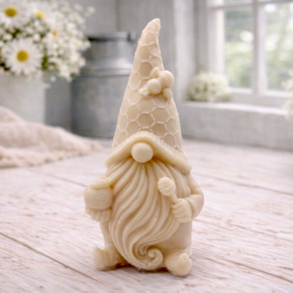 A handmade vegan soap in the shape of a gnome with a long beard, wearing a honeycomb-patterned hat with a bee, holding a honey pot and dipper.