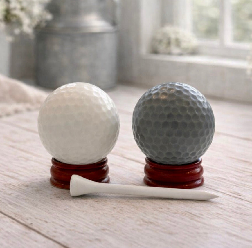 A handmade vegan golf ball soap gift set featuring one white and one grey spherical soap with realistic dimpled textures, displayed on small wooden stands next to a white golf tee and a kraft gift box tied with a red ribbon