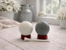 Load image into Gallery viewer, A handmade vegan golf ball soap gift set featuring one white and one grey spherical soap with realistic dimpled textures, displayed on small wooden stands next to a white golf tee and a kraft gift box tied with a red ribbon
