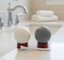 Load image into Gallery viewer, A set of two handmade vegan golf ball soaps in white and grey, displayed on miniature wooden stands with a golf tee in a bright farmhouse decor bathroom.

