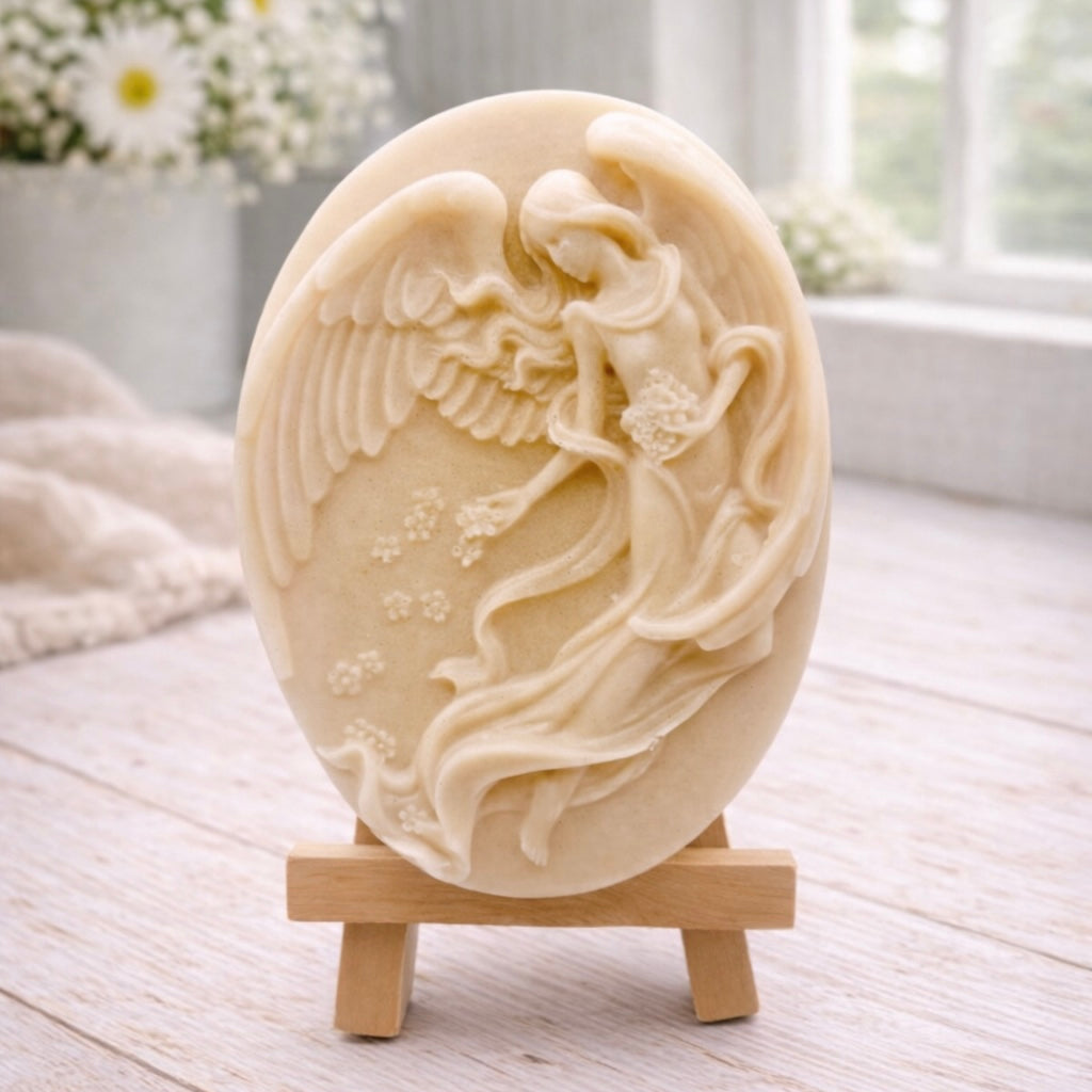 An oval-shaped handmade vegan soap, featuring an intricate relief carving of a guardian angel with large wings holding flowers, displayed on a small wooden easel.