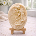 An oval-shaped handmade vegan soap, featuring an intricate relief carving of a guardian angel with large wings holding flowers, displayed on a small wooden easel.