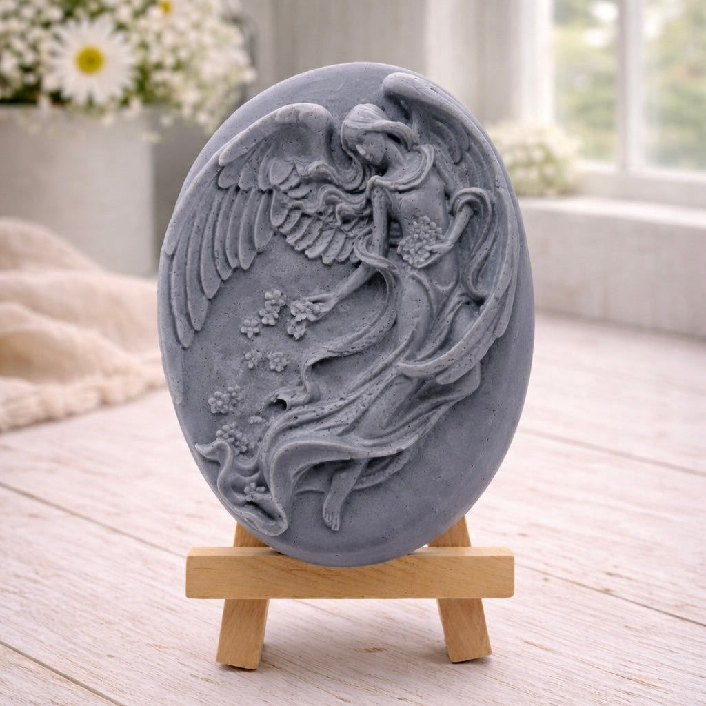 An oval-shaped handmade vegan soap, featuring an intricate relief carving of a guardian angel with large wings holding flowers, displayed on a small wooden easel.