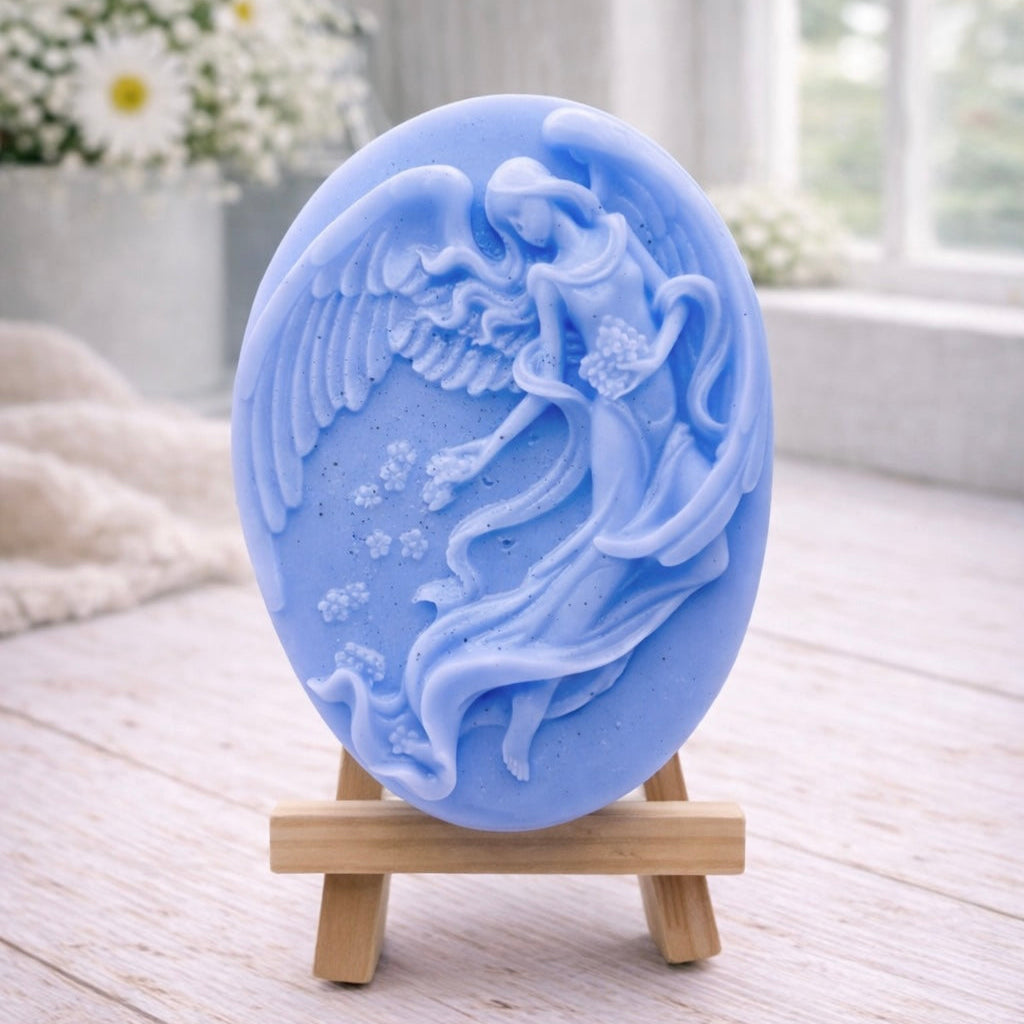 An oval-shaped handmade vegan soap, featuring an intricate relief carving of a guardian angel with large wings holding flowers, displayed on a small wooden easel.
