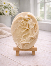 Load image into Gallery viewer, An oval-shaped handmade vegan soap, featuring an intricate relief carving of a guardian angel with large wings holding flowers, displayed on a small wooden easel.
