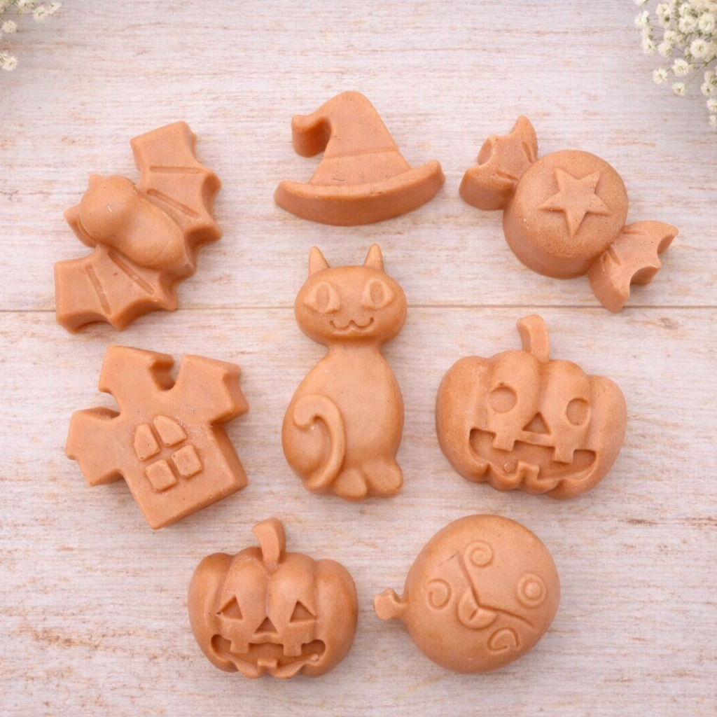 A collection of eight handmade vegan Halloween soaps featuring a bat, witch's hat, star-patterned sweet, haunted house, black cat, jack-o'-lantern, and a ghost, arranged on a light wooden surface with white floral accents.