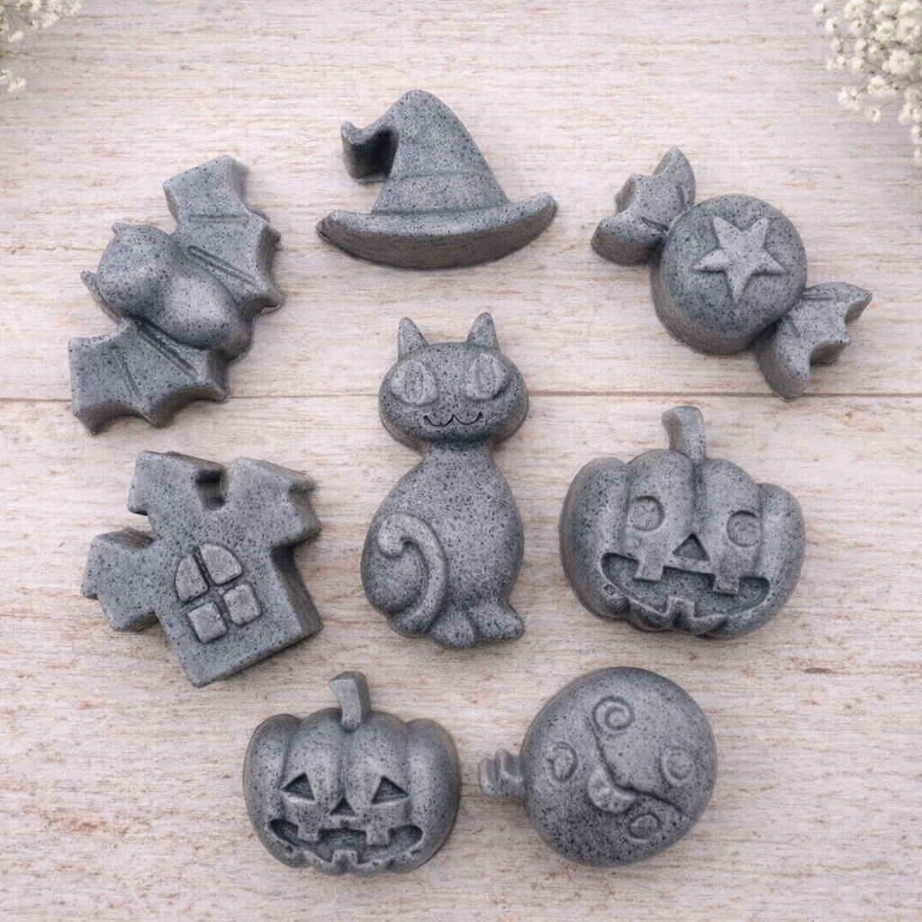 A collection of eight handmade vegan Halloween soaps featuring a bat, witch's hat, star-patterned sweet, haunted house, black cat, jack-o'-lantern, and a ghost, arranged on a light wooden surface with white floral accents.