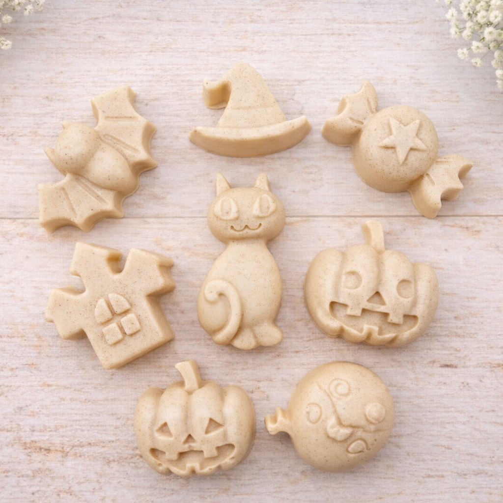 A collection of eight handmade vegan Halloween soaps featuring a bat, witch's hat, star-patterned sweet, haunted house, black cat, jack-o'-lantern, and a ghost, arranged on a light wooden surface with white floral accents.