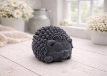 Load image into Gallery viewer, A detailed handmade vegan soap sculpted in the shape of a round, sitting hedgehog with intricate quill textures and a cute face, in a natural honey-brown tone, displayed on a light wooden surface.
