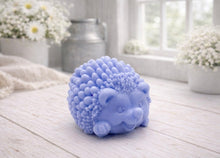 Load image into Gallery viewer, A detailed handmade vegan soap sculpted in the shape of a round, sitting hedgehog with intricate quill textures and a cute face, in a natural honey-brown tone, displayed on a light wooden surface.
