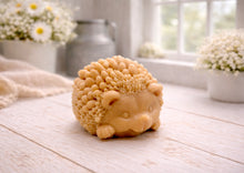 Load image into Gallery viewer, A detailed handmade vegan soap sculpted in the shape of a round, sitting hedgehog with intricate quill textures and a cute face, in a natural honey-brown tone, displayed on a light wooden surface.
