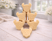 Load image into Gallery viewer, A collection of six handmade vegan soaps each intricately sculpted into the head of a shaggy Highland cow with long horns and a thick, textured coat, arranged in a pyramid on and around a small wooden easel.
