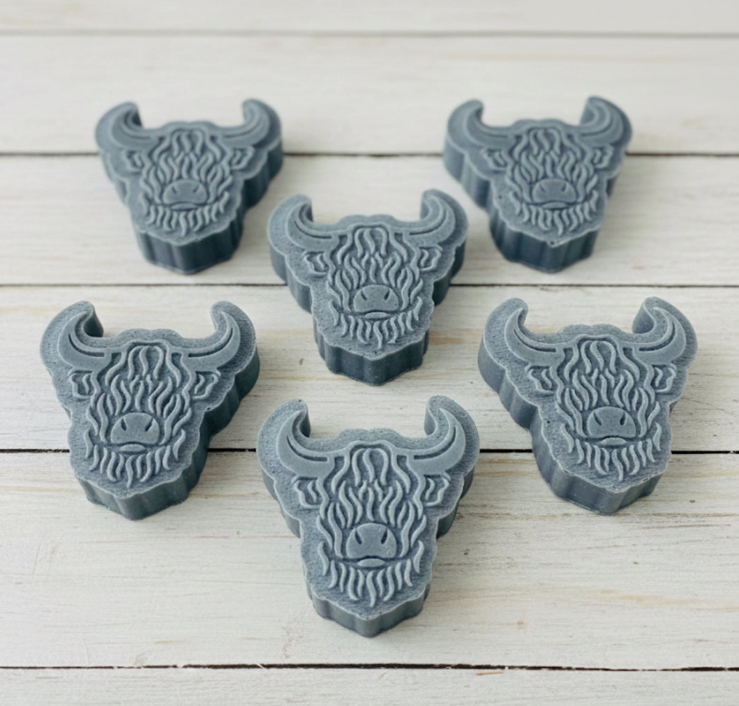 A collection of six handmade vegan soaps each intricately sculpted into the head of a shaggy Highland cow with long horns and a thick, textured coat, arranged in a pyramid on and around a small wooden easel.