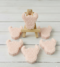 Load image into Gallery viewer, A collection of six handmade vegan soaps each intricately sculpted into the head of a shaggy Highland cow with long horns and a thick, textured coat, arranged in a pyramid on and around a small wooden easel.
