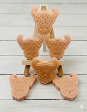 Load image into Gallery viewer, A collection of six handmade vegan soaps each intricately sculpted into the head of a shaggy Highland cow with long horns and a thick, textured coat, arranged in a pyramid on and around a small wooden easel.
