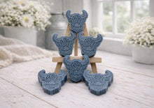 Load image into Gallery viewer, A collection of six handmade vegan soaps each intricately sculpted into the head of a shaggy Highland cow with long horns and a thick, textured coat, arranged in a pyramid on and around a small wooden easel.
