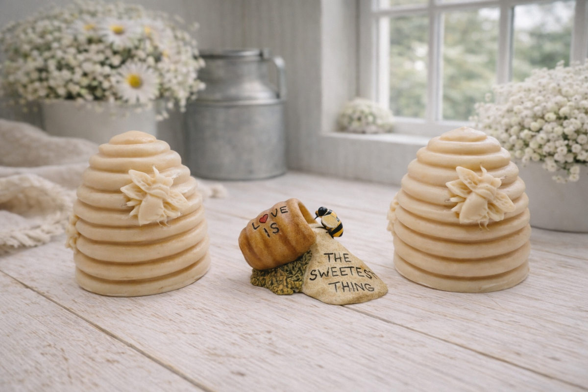 Two handmade vegan soaps sculpted into traditional dome-shaped beehives (skeps) with small bees resting on the sides, flanking a decorative centrepiece featuring a honey pot and the words "The Sweetest Thing," all displayed on a rustic wooden surface.