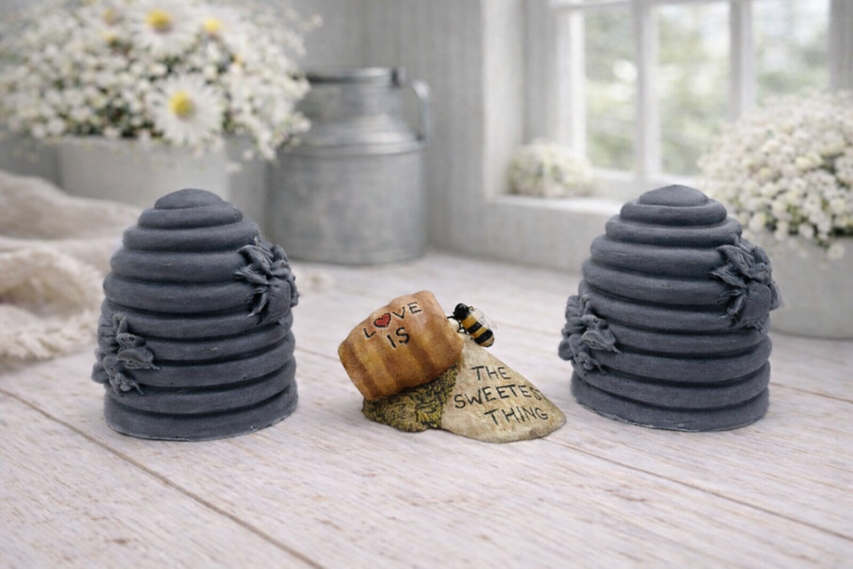 Two handmade vegan soaps sculpted into traditional dome-shaped beehives (skeps) with small bees resting on the sides, flanking a decorative centrepiece featuring a honey pot and the words "The Sweetest Thing," all displayed on a rustic wooden surface.
