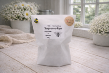 Load image into Gallery viewer, Bag of soap-on-a-rope with bee illustrations on a wooden surface with flowers in the background
