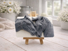 Load image into Gallery viewer, A rectangular handmade vegan soap featuring a detailed relief of two horse heads leaning affectionately towards each other, finished in a slate-grey tone against a white soap base and displayed on a miniature wooden easel.
