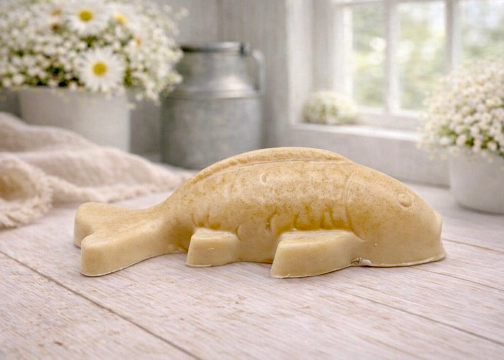 Handmade vegan soap sculpted in the shape of a koi carp fish with detailed scales and fins, resting on a light timber surface with a soft floral background.