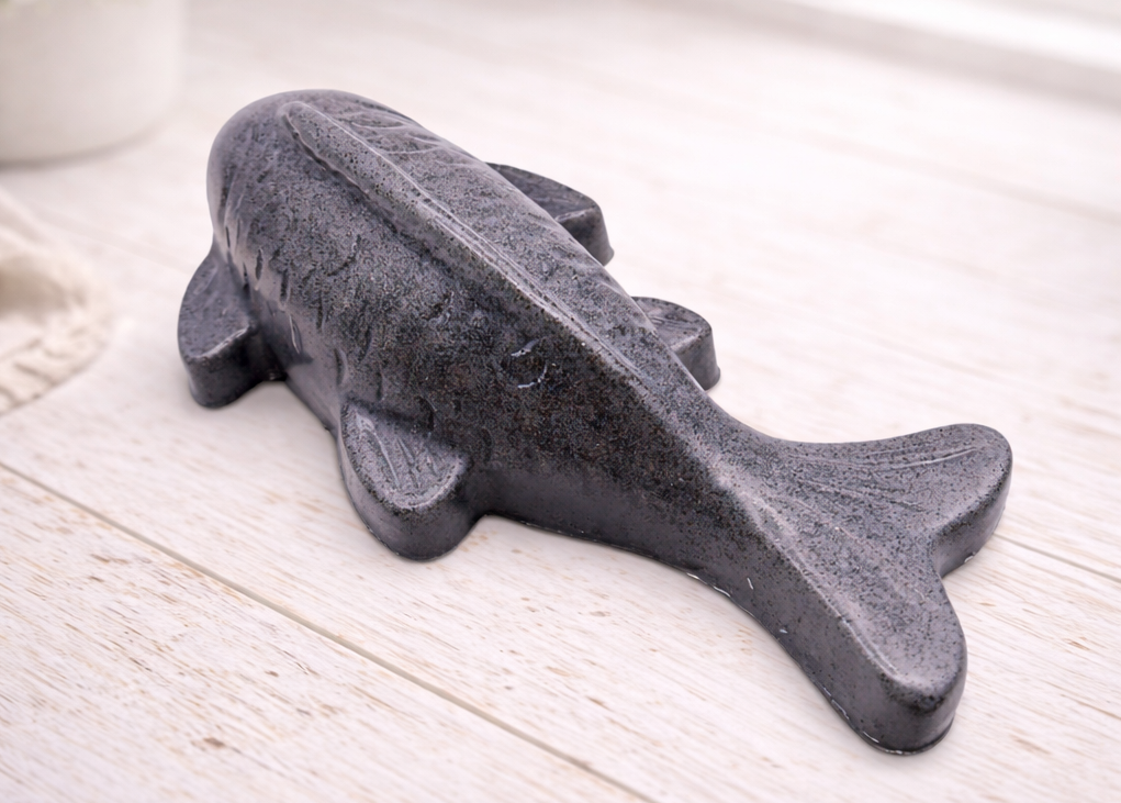 Handmade vegan soap sculpted in the shape of a koi carp fish with detailed scales and fins, resting on a light timber surface with a soft floral background.