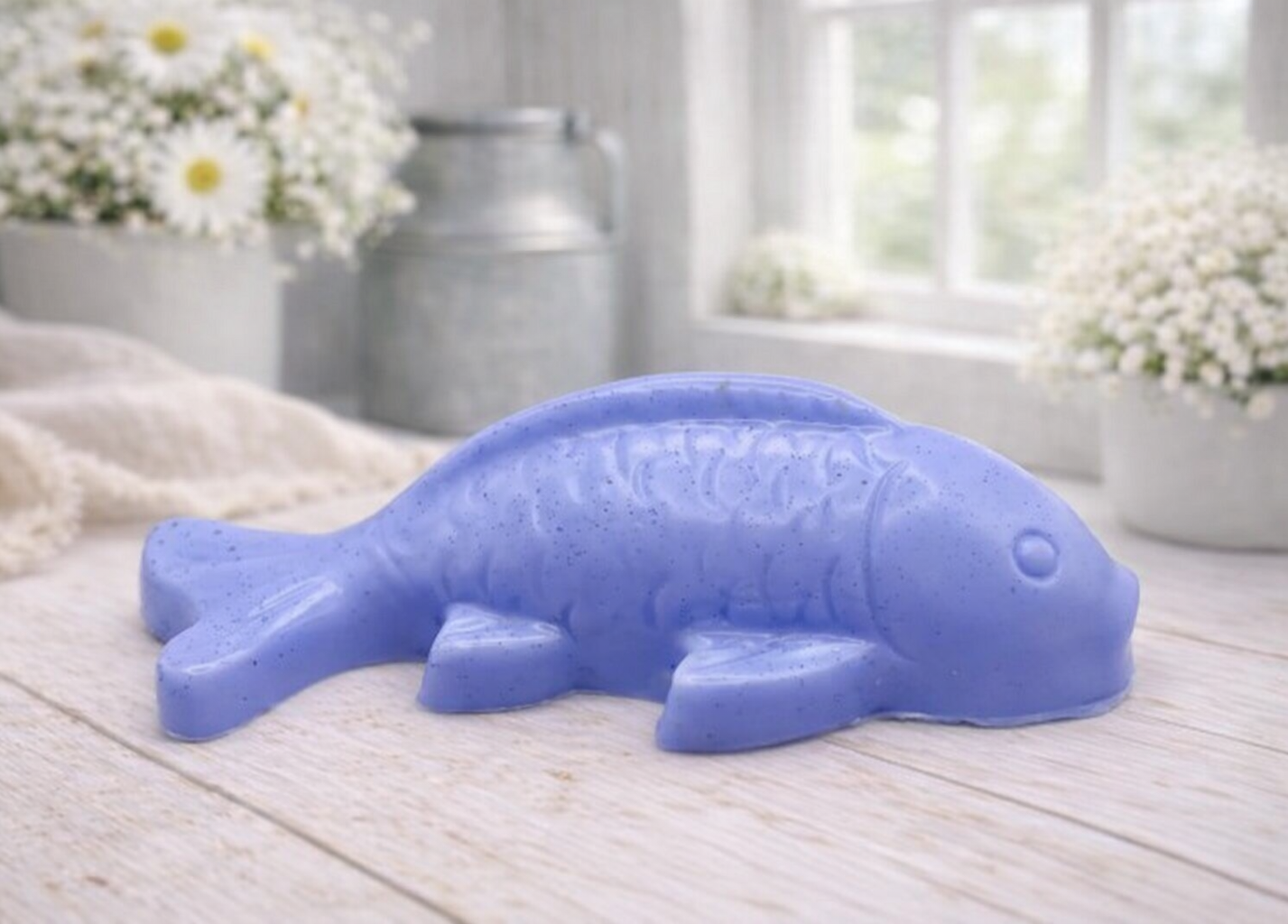 Handmade vegan soap sculpted in the shape of a koi carp fish with detailed scales and fins, resting on a light timber surface with a soft floral background.