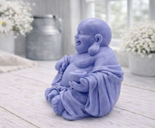 Load image into Gallery viewer, A 3D handmade vegan soap sculpted into a joyful Laughing Buddha sitting in a meditative pose, holding prayer beads and a small bowl, displayed in a bright farmhouse setting
