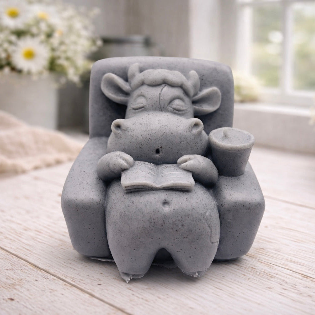 A handmade vegan soap sculpted to show a whimsical "lazy cow" character relaxing in an armchair, reading a book and holding a drink, displayed on a light wooden surface.