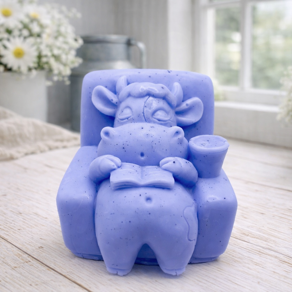 A handmade vegan soap sculpted to show a whimsical "lazy cow" character relaxing in an armchair, reading a book and holding a drink, displayed on a light wooden surface.