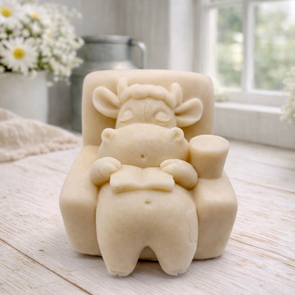 A handmade vegan soap sculpted to show a whimsical "lazy cow" character relaxing in an armchair, reading a book and holding a drink, displayed on a light wooden surface.