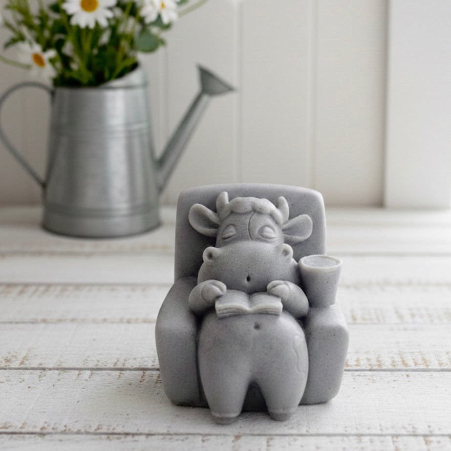 A handmade vegan soap sculpted to show a whimsical "lazy cow" character relaxing in an armchair, reading a book and holding a drink, displayed on a light wooden surface.