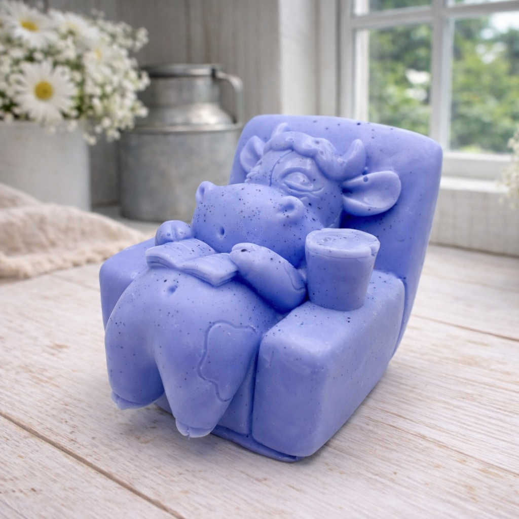 A handmade vegan soap sculpted to show a whimsical "lazy cow" character relaxing in an armchair, reading a book and holding a drink, displayed on a light wooden surface.