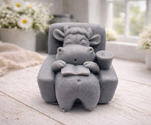 Load image into Gallery viewer, A handmade vegan soap sculpted to show a whimsical "lazy cow" character relaxing in an armchair, reading a book and holding a drink, displayed on a light wooden surface.
