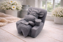 Load image into Gallery viewer, A handmade vegan soap sculpted to show a whimsical "lazy cow" character relaxing in an armchair, reading a book and holding a drink, displayed on a light wooden surface.
