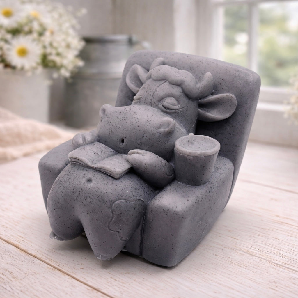 A handmade vegan soap sculpted to show a whimsical "lazy cow" character relaxing in an armchair, reading a book and holding a drink, displayed on a light wooden surface.