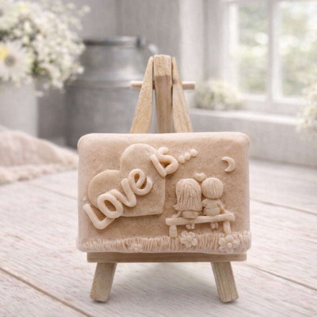 A rectangular handmade vegan soap featuring a detailed 3D relief carving of a young couple sitting on a bench under a crescent moon, with the words "Love is..." embossed inside a heart, displayed on a wooden easel.