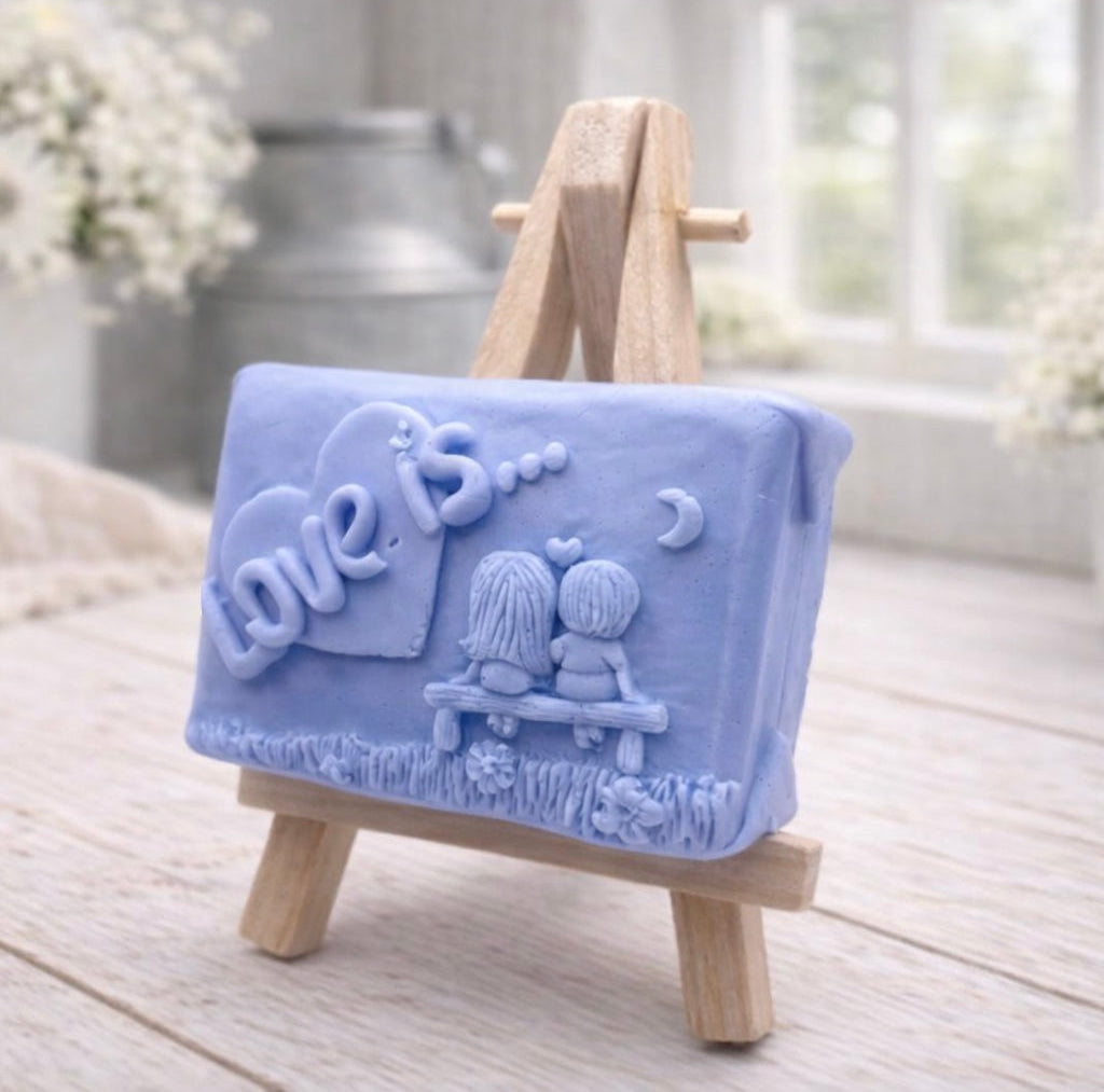 A rectangular handmade vegan soap featuring a detailed 3D relief carving of a young couple sitting on a bench under a crescent moon, with the words "Love is..." embossed inside a heart, displayed on a wooden easel.