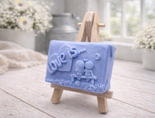 Load image into Gallery viewer, A rectangular handmade vegan soap featuring a detailed 3D relief carving of a young couple sitting on a bench under a crescent moon, with the words "Love is..." embossed inside a heart, displayed on a wooden easel.
