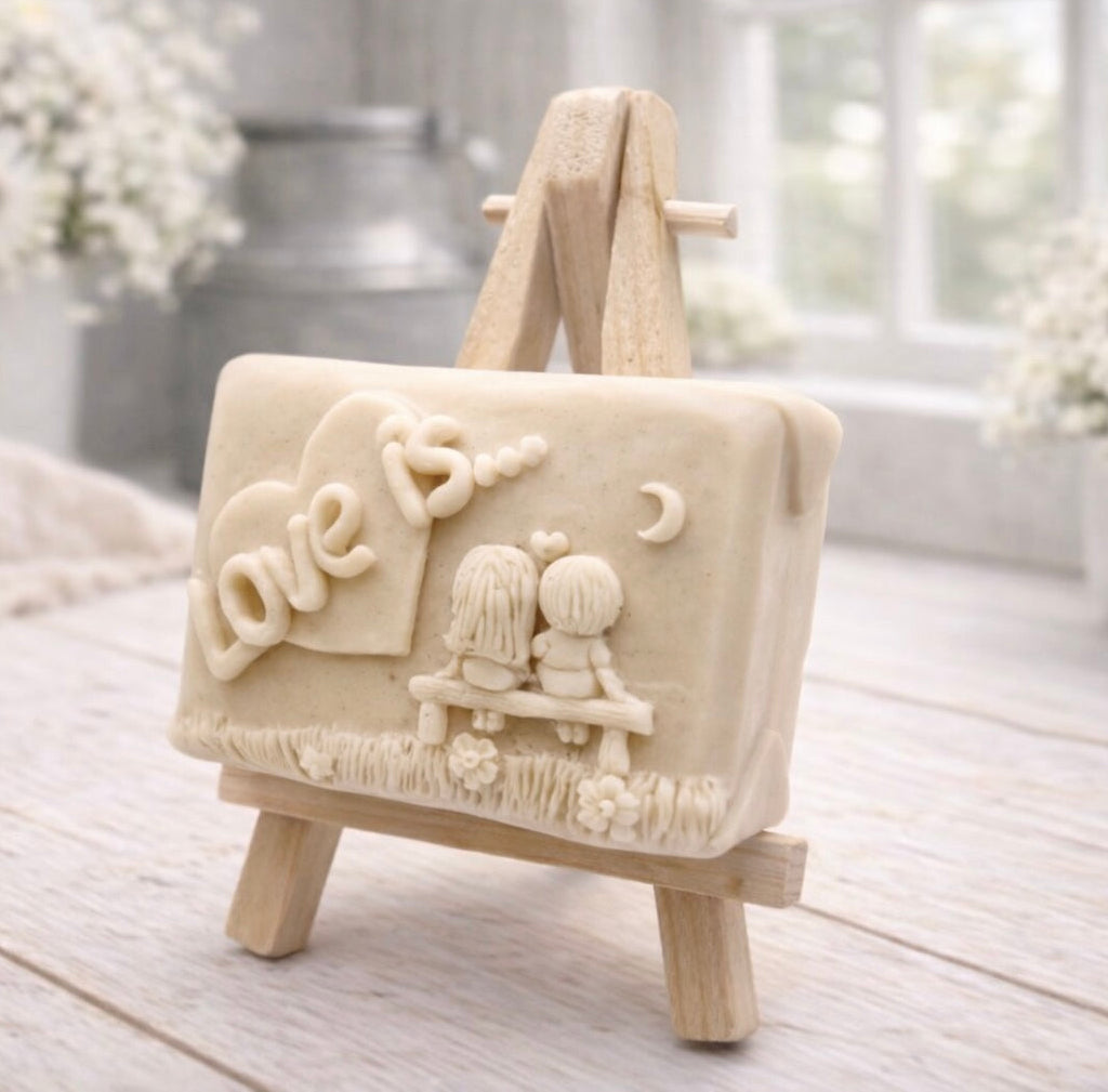 A rectangular handmade vegan soap featuring a detailed 3D relief carving of a young couple sitting on a bench under a crescent moon, with the words "Love is..." embossed inside a heart, displayed on a wooden easel.