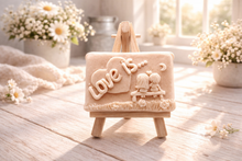 Load image into Gallery viewer, A rectangular handmade vegan soap featuring a detailed 3D relief carving of a young couple sitting on a bench under a crescent moon, with the words "Love is..." embossed inside a heart, displayed on a wooden easel.
