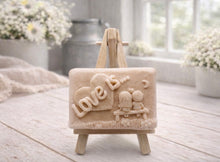 Load image into Gallery viewer, A rectangular handmade vegan soap featuring a detailed 3D relief carving of a young couple sitting on a bench under a crescent moon, with the words "Love is..." embossed inside a heart, displayed on a wooden easel.

